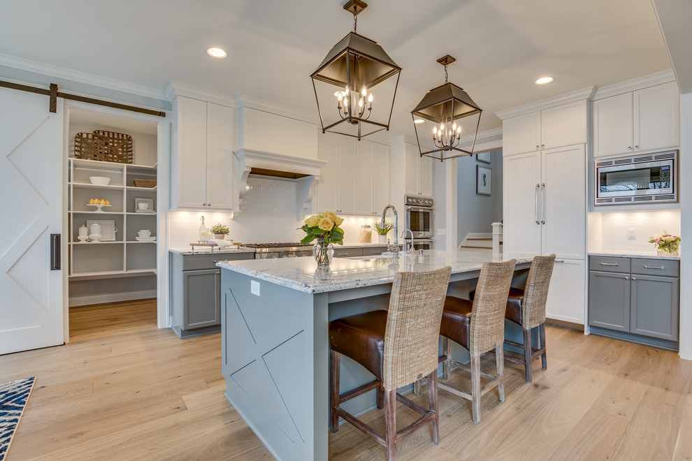 This is an example of a large country galley kitchen pantry in Minneapolis with a belfast sink, recessed-panel cabinets, white cabinets, white splashback, metro tiled splashback, stainless steel appliances, light hardwood flooring, an island and beige floors.