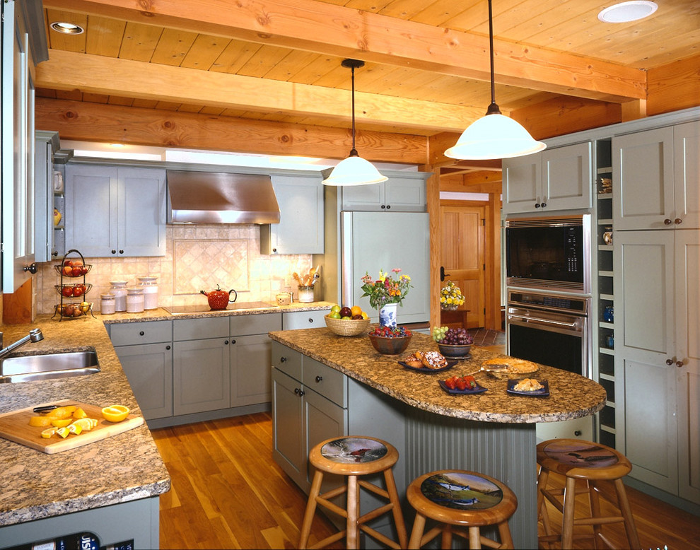 Cette image montre une cuisine américaine traditionnelle en U avec un évier encastré, un placard avec porte à panneau encastré, des portes de placard bleues, une crédence beige, un électroménager en acier inoxydable, un sol en bois brun et îlot.