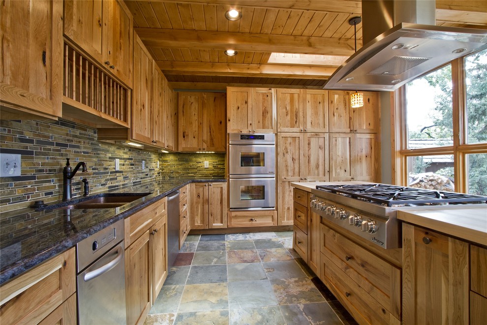 This is an example of a rustic l-shaped kitchen/diner in Denver with a double-bowl sink, recessed-panel cabinets, light wood cabinets, granite worktops, green splashback, stone tiled splashback, stainless steel appliances, slate flooring and an island.