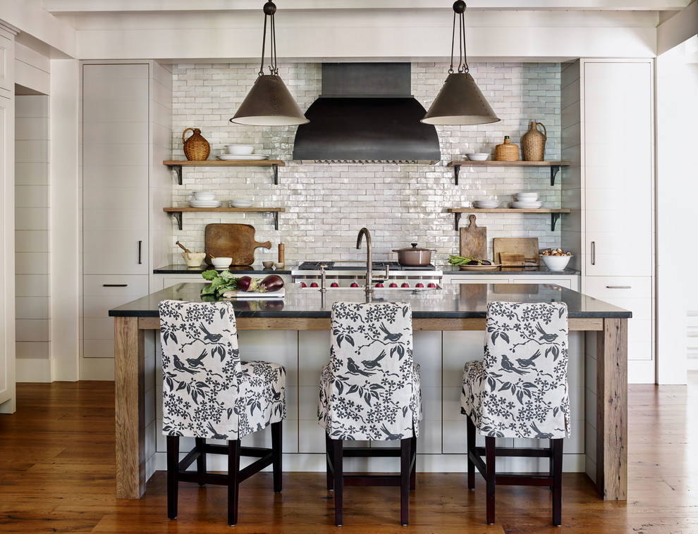 Example of a mountain style galley medium tone wood floor and brown floor kitchen design with a farmhouse sink, flat-panel cabinets, gray cabinets, white backsplash, subway tile backsplash, stainless steel appliances, an island and black countertops