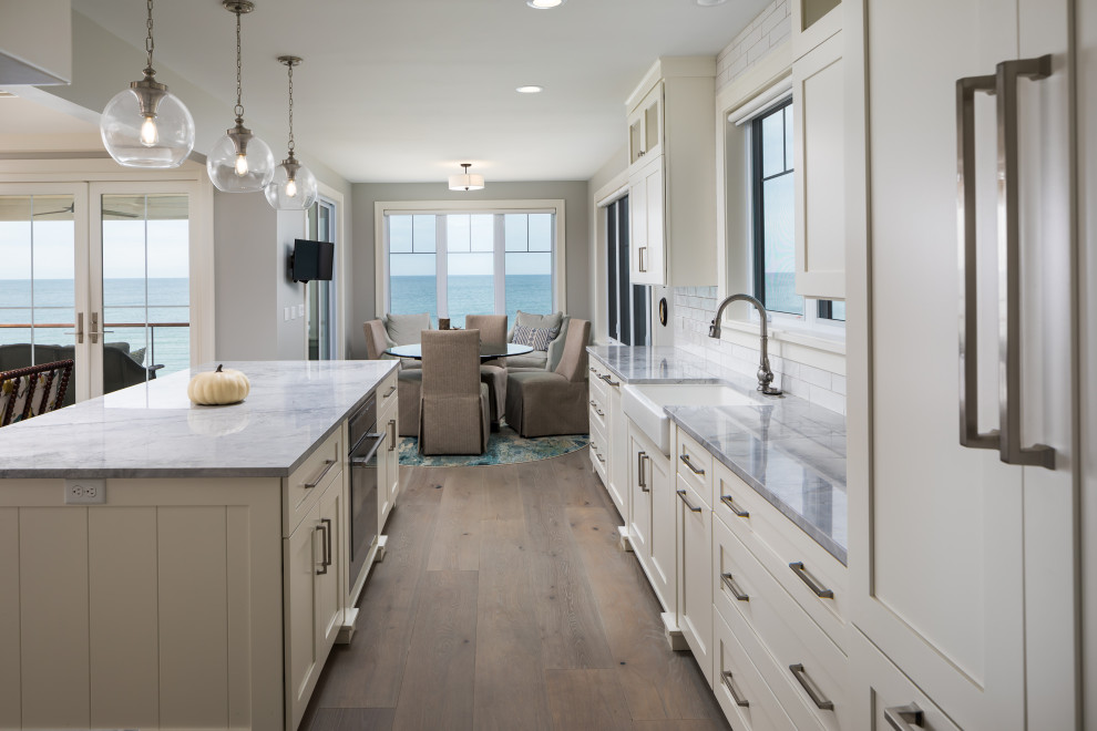 Inspiration for a medium sized traditional l-shaped open plan kitchen in Grand Rapids with a belfast sink, recessed-panel cabinets, white cabinets, quartz worktops, white splashback, metro tiled splashback, stainless steel appliances, light hardwood flooring, an island, grey floors and grey worktops.