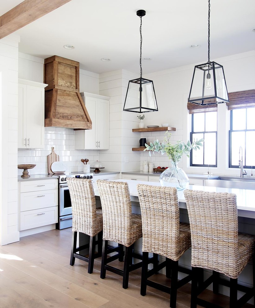 Photo of a farmhouse l-shaped kitchen in Other with a belfast sink, shaker cabinets, white cabinets, white splashback, stainless steel appliances, light hardwood flooring, an island, beige floors and white worktops.