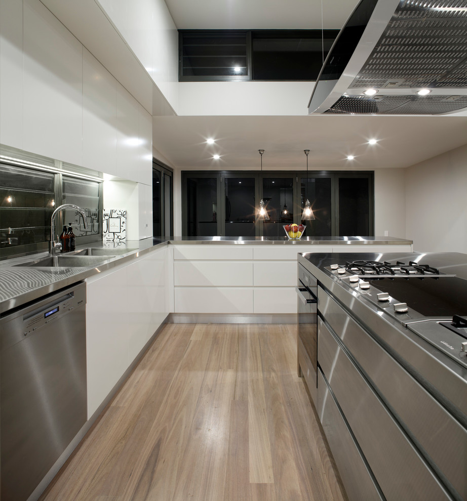This is an example of a large contemporary l-shaped open plan kitchen in Sydney with a submerged sink, flat-panel cabinets, white cabinets, stainless steel worktops, white splashback, mosaic tiled splashback, stainless steel appliances, light hardwood flooring and an island.