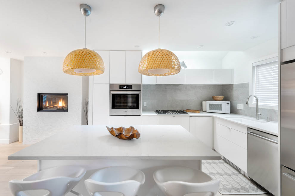 Bild på ett funkis vit vitt l-kök, med en undermonterad diskho, luckor med glaspanel, vita skåp, grått stänkskydd, stänkskydd i cementkakel, rostfria vitvaror, ljust trägolv och en köksö