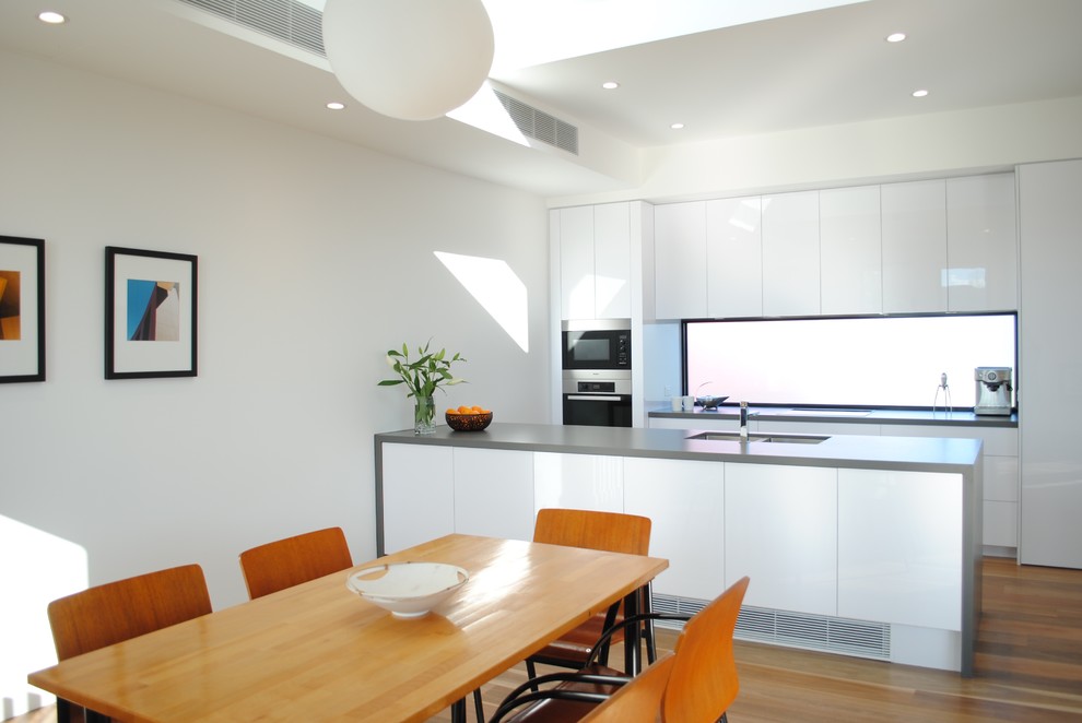 Photo of a small contemporary single-wall open plan kitchen in Melbourne with a submerged sink, flat-panel cabinets, white cabinets, engineered stone countertops, stainless steel appliances, an island, window splashback and medium hardwood flooring.