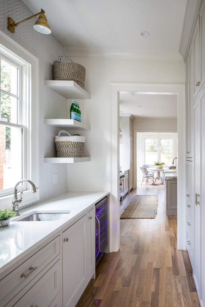Inspiration for a medium sized classic kitchen pantry in Atlanta with a belfast sink, beaded cabinets, white cabinets, engineered stone countertops, white splashback, porcelain splashback, integrated appliances, light hardwood flooring, an island, brown floors and white worktops.
