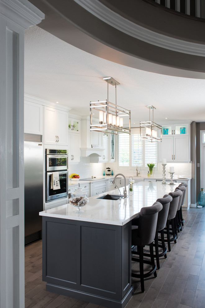 Example of a large transitional l-shaped medium tone wood floor eat-in kitchen design in Toronto with a farmhouse sink, recessed-panel cabinets, white cabinets, quartz countertops, white backsplash, ceramic backsplash, stainless steel appliances and an island
