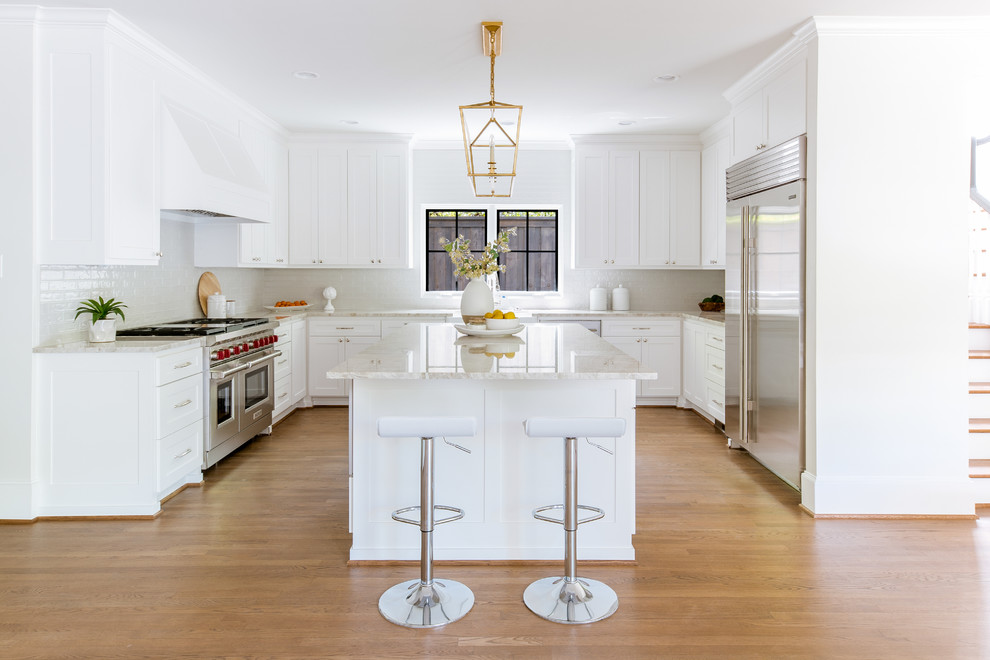 Example of a transitional u-shaped medium tone wood floor and brown floor kitchen design in Dallas with a farmhouse sink, shaker cabinets, white cabinets, white backsplash, subway tile backsplash, stainless steel appliances, an island and beige countertops