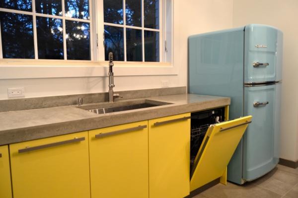На фото: угловая кухня среднего размера в стиле модернизм с обеденным столом, накладной мойкой, плоскими фасадами, желтыми фасадами, столешницей из акрилового камня, цветной техникой, полом из керамической плитки и островом