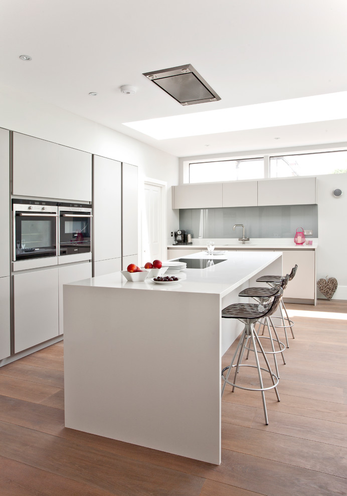 Moderne Küche in L-Form mit Unterbauwaschbecken, flächenbündigen Schrankfronten, weißen Schränken, Küchenrückwand in Grau, Glasrückwand, Elektrogeräten mit Frontblende, braunem Holzboden und Kücheninsel in Surrey