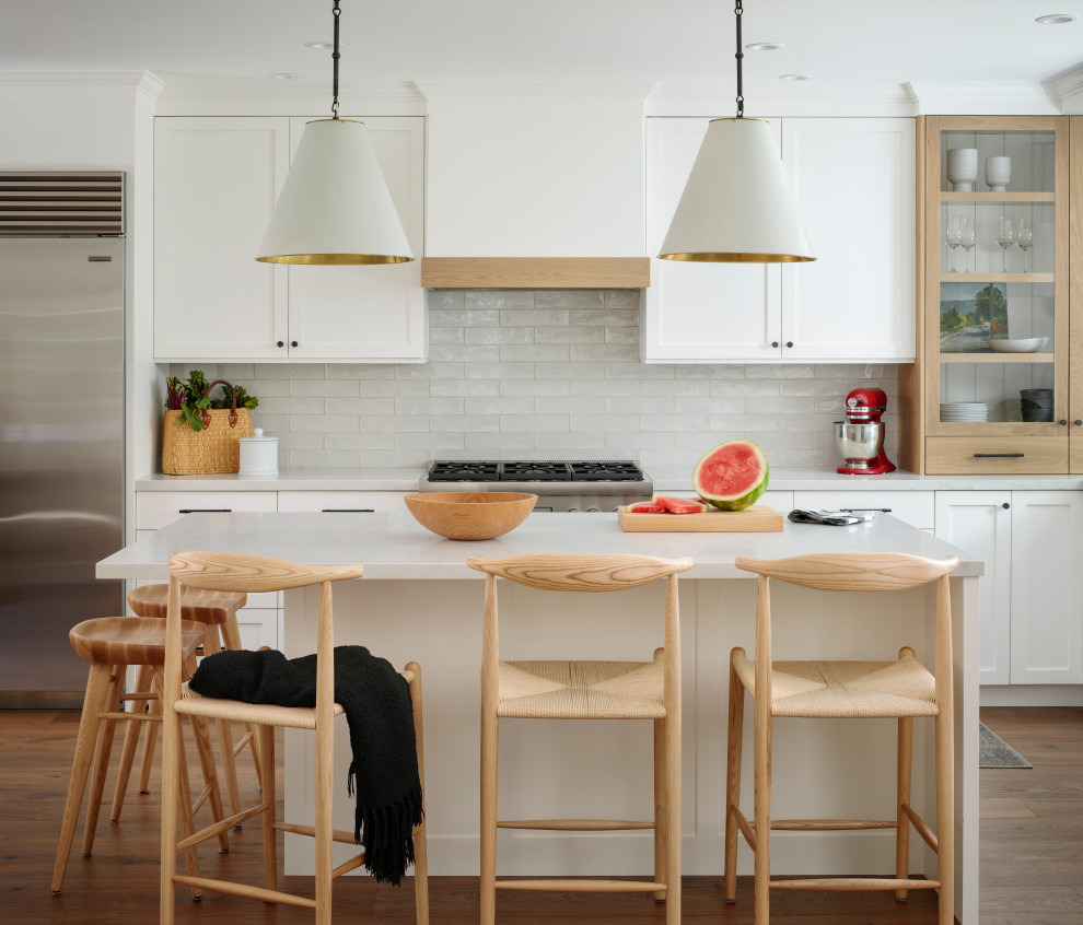 Bild på ett mellanstort vintage vit vitt l-kök, med en undermonterad diskho, skåp i shakerstil, vita skåp, bänkskiva i kvarts, grått stänkskydd, stänkskydd i porslinskakel, rostfria vitvaror, mellanmörkt trägolv, en köksö och brunt golv