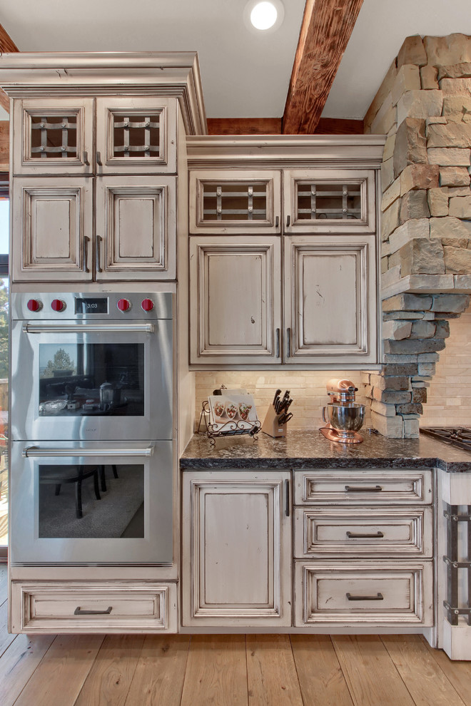 Bild på ett mycket stort rustikt grå grått kök, med en rustik diskho, luckor med upphöjd panel, vita skåp, granitbänkskiva, beige stänkskydd, stänkskydd i travertin, integrerade vitvaror, ljust trägolv, en köksö och beiget golv