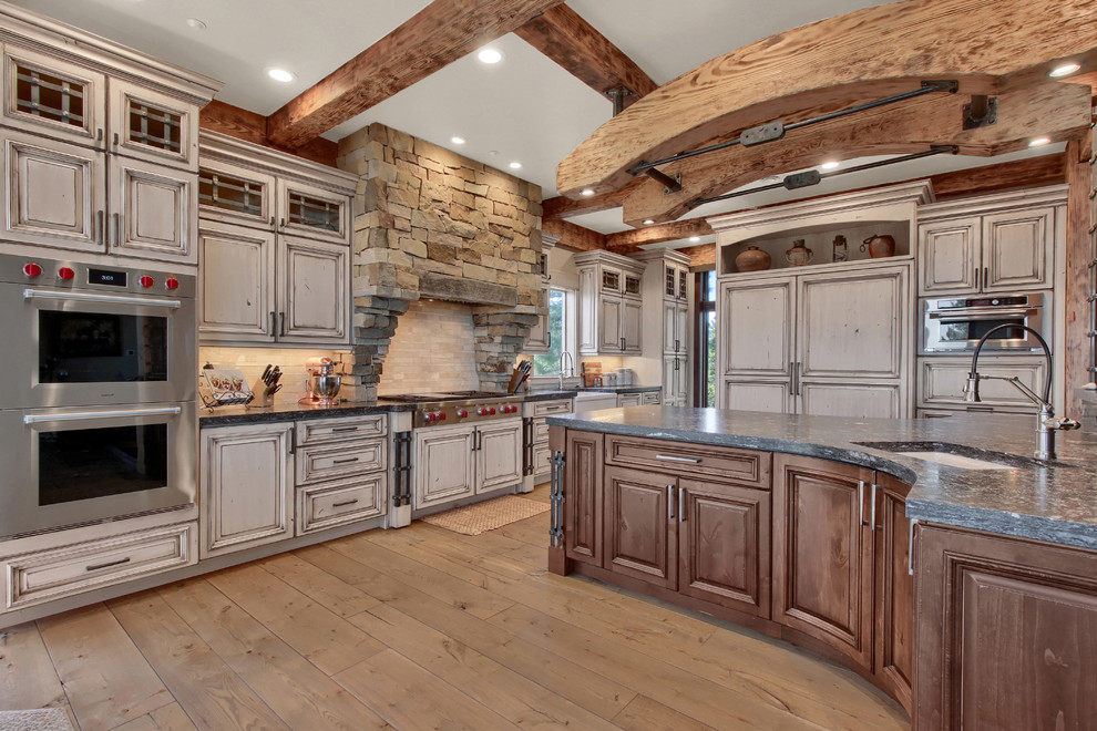 Inredning av ett rustikt mycket stort grå grått kök, med en rustik diskho, luckor med upphöjd panel, granitbänkskiva, beige stänkskydd, stänkskydd i travertin, integrerade vitvaror, en köksö, grå skåp, mellanmörkt trägolv och brunt golv