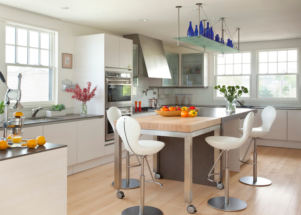 Large contemporary u-shaped enclosed kitchen in Boston with flat-panel cabinets, white cabinets, wood worktops, a submerged sink, stainless steel appliances, light hardwood flooring, multiple islands, white splashback, glass sheet splashback and brown floors.