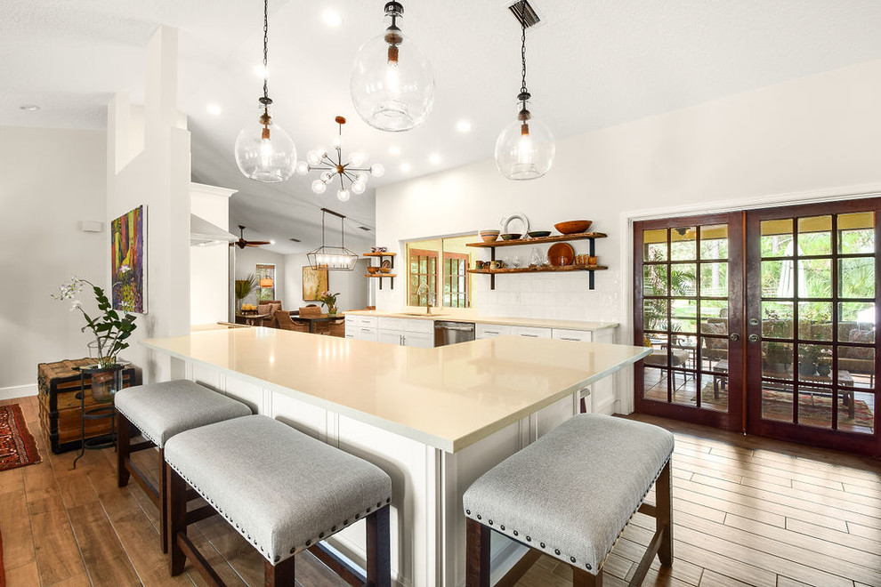 This is an example of a medium sized contemporary galley kitchen/diner in Miami with a triple-bowl sink, recessed-panel cabinets, white cabinets, engineered stone countertops, white splashback, porcelain splashback, stainless steel appliances, laminate floors, a breakfast bar, brown floors and white worktops.