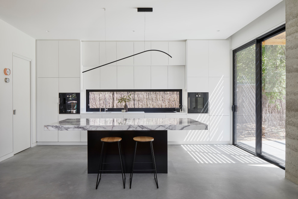 Mid-sized trendy galley concrete floor and gray floor open concept kitchen photo in Melbourne with an undermount sink, flat-panel cabinets, white cabinets, marble countertops, black appliances, an island and white countertops