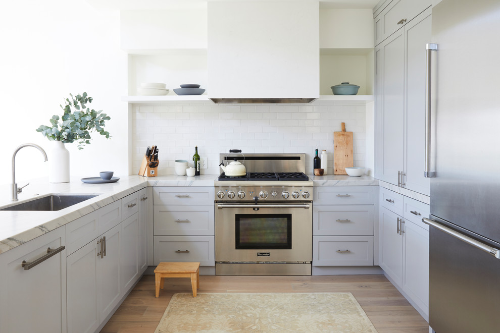 Traditional u-shaped kitchen in San Francisco with a submerged sink, recessed-panel cabinets, grey cabinets, white splashback, stainless steel appliances, light hardwood flooring, a breakfast bar and beige floors.