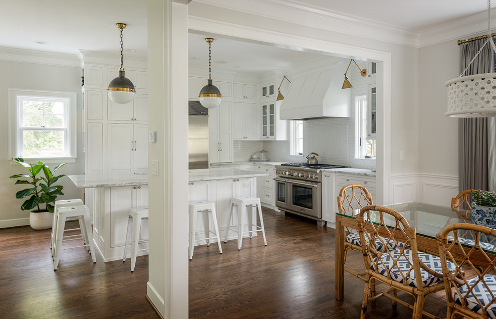 Exemple d'une cuisine américaine chic en L avec un placard à porte shaker, des portes de placard blanches, une crédence blanche, une crédence en carrelage métro, un électroménager en acier inoxydable, parquet foncé et îlot.