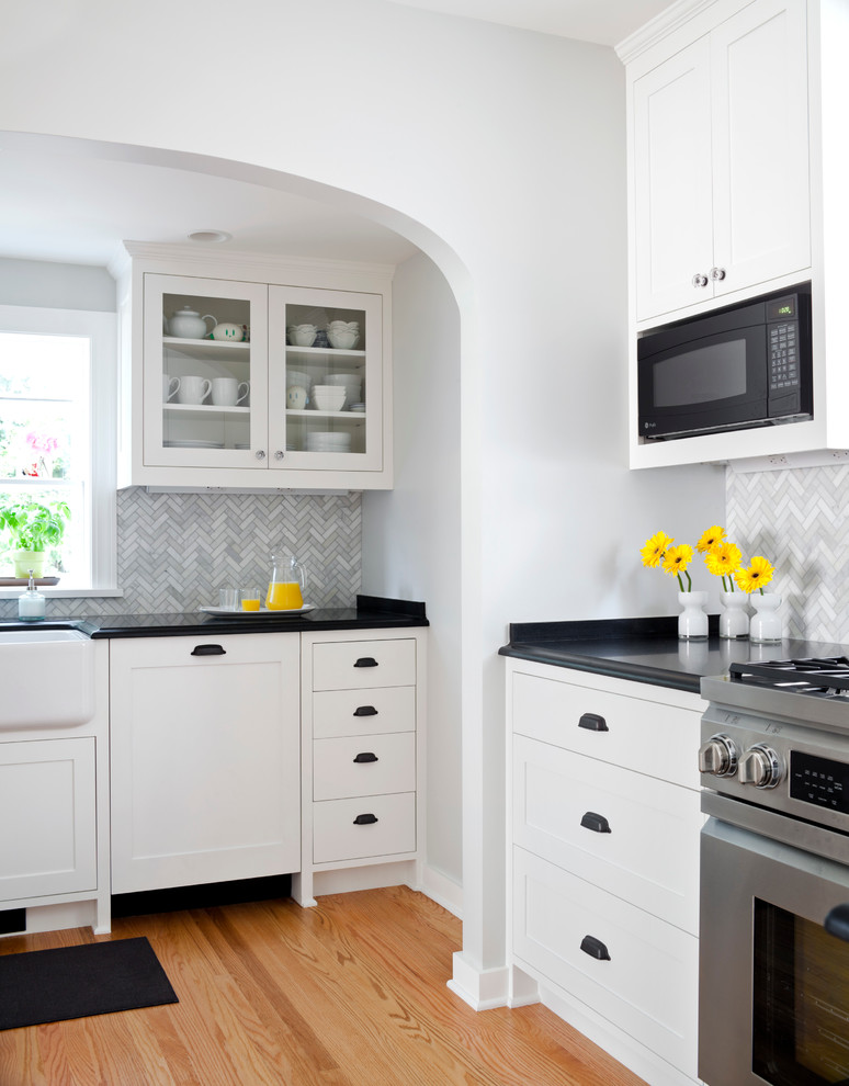 Klassisk inredning av ett stort kök, med en rustik diskho, luckor med infälld panel, vita skåp, granitbänkskiva, grått stänkskydd, stänkskydd i glaskakel, rostfria vitvaror och ljust trägolv