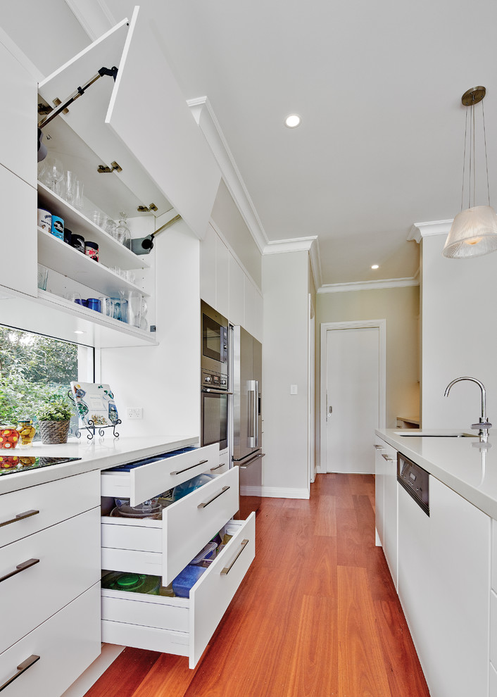 Inredning av ett modernt stort kök, med en integrerad diskho, släta luckor, vita skåp, bänkskiva i kvarts, fönster som stänkskydd, rostfria vitvaror, mellanmörkt trägolv, en köksö och brunt golv