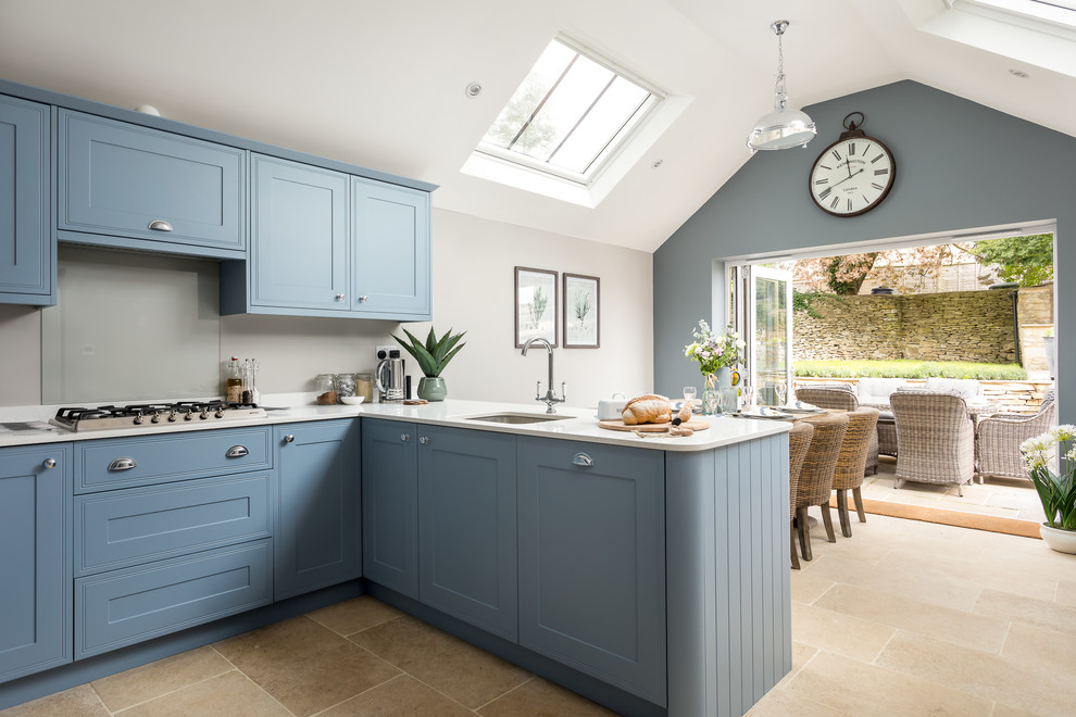 Große Country Wohnküche in L-Form mit Waschbecken, blauen Schränken, beigem Boden, Kassettenfronten, Küchenrückwand in Grau, Glasrückwand, Halbinsel und weißer Arbeitsplatte in Gloucestershire