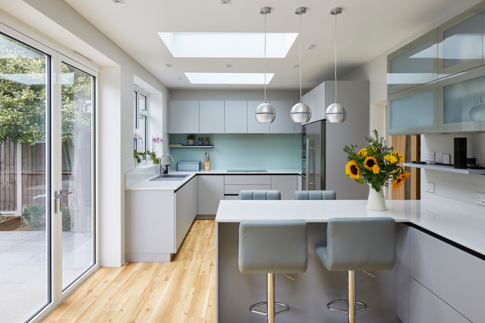 Example of a mid-sized trendy u-shaped eat-in kitchen design in London with flat-panel cabinets, gray cabinets, quartzite countertops, green backsplash, glass sheet backsplash, stainless steel appliances, a peninsula and white countertops