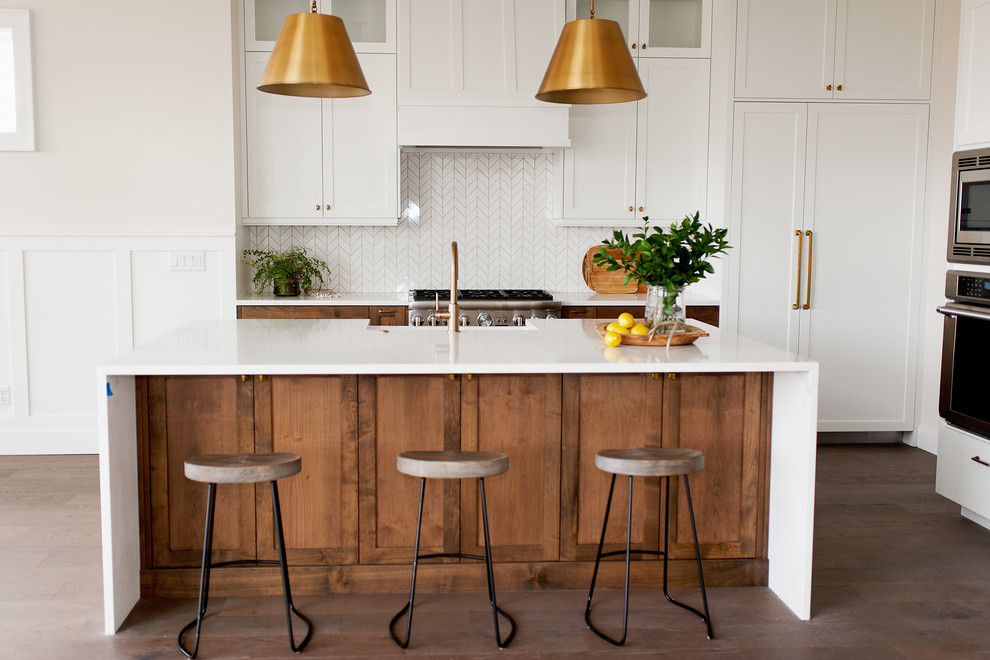 Inspiration for a medium sized rural l-shaped kitchen in Boise with a belfast sink, shaker cabinets, medium wood cabinets, engineered stone countertops, white splashback, mosaic tiled splashback, stainless steel appliances, medium hardwood flooring, an island, brown floors and white worktops.
