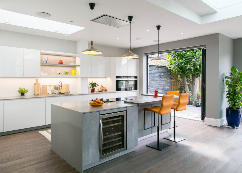 Foto på ett stort funkis grå kök, med en köksö, en undermonterad diskho, släta luckor, vita skåp, vitt stänkskydd, rostfria vitvaror, mellanmörkt trägolv och brunt golv