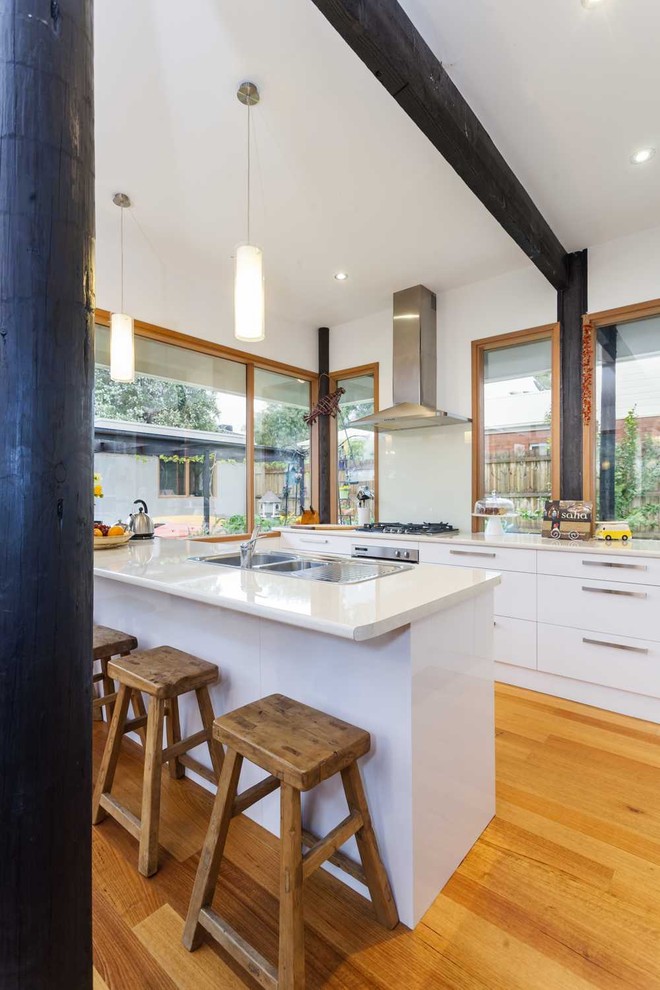 Example of a mid-sized 1960s u-shaped medium tone wood floor and beige floor eat-in kitchen design in Melbourne with flat-panel cabinets, white cabinets, a double-bowl sink, stainless steel appliances, an island, white backsplash, glass sheet backsplash and white countertops