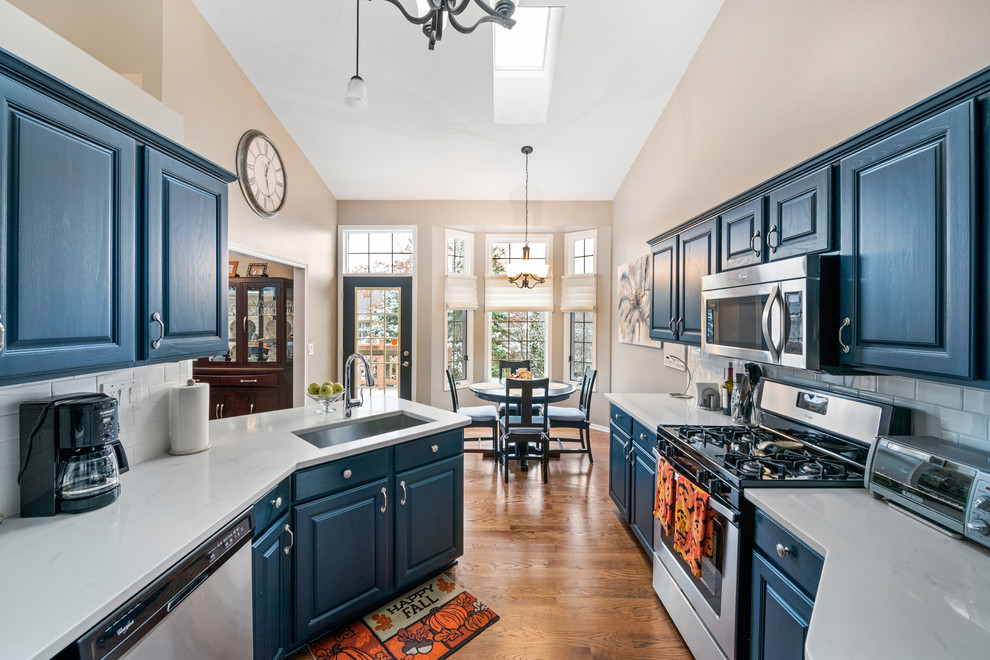 Bild på ett mellanstort vintage vit vitt kök, med en enkel diskho, luckor med upphöjd panel, blå skåp, bänkskiva i kvarts, vitt stänkskydd, stänkskydd i cementkakel, rostfria vitvaror, mellanmörkt trägolv, en halv köksö och brunt golv