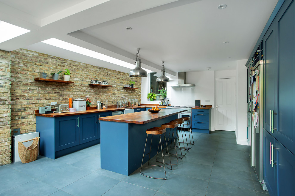 ロンドンにある高級な広いトランジショナルスタイルのおしゃれなキッチン (エプロンフロントシンク、シェーカースタイル扉のキャビネット、青いキャビネット、木材カウンター、シルバーの調理設備、セラミックタイルの床、グレーの床、茶色いキッチンカウンター、茶色いキッチンパネル、レンガのキッチンパネル) の写真