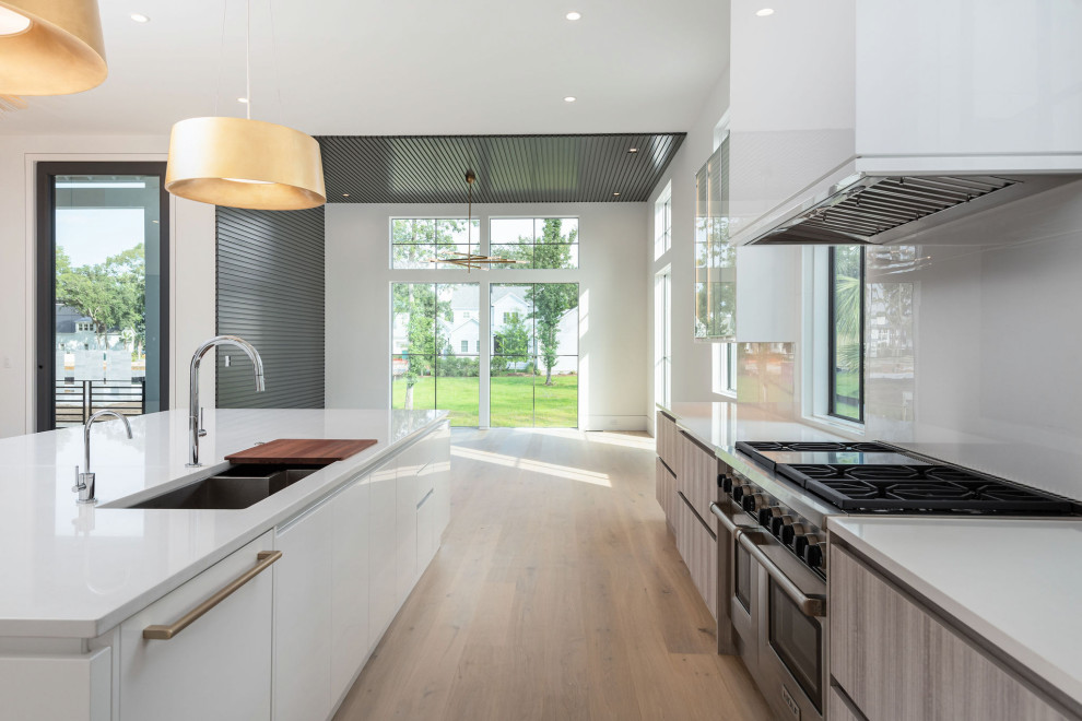Photo of a large modern l-shaped kitchen/diner in Charleston with a double-bowl sink, recessed-panel cabinets, white cabinets, engineered stone countertops, white splashback, porcelain splashback, integrated appliances, light hardwood flooring, an island, beige floors and white worktops.