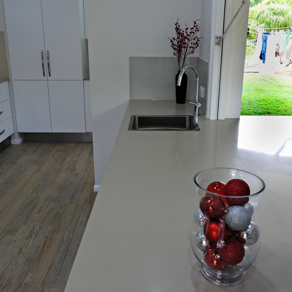 Photo of a medium sized contemporary galley kitchen pantry in Brisbane with a single-bowl sink, flat-panel cabinets, white cabinets, engineered stone countertops, beige splashback, glass sheet splashback, stainless steel appliances, vinyl flooring, a breakfast bar and brown floors.