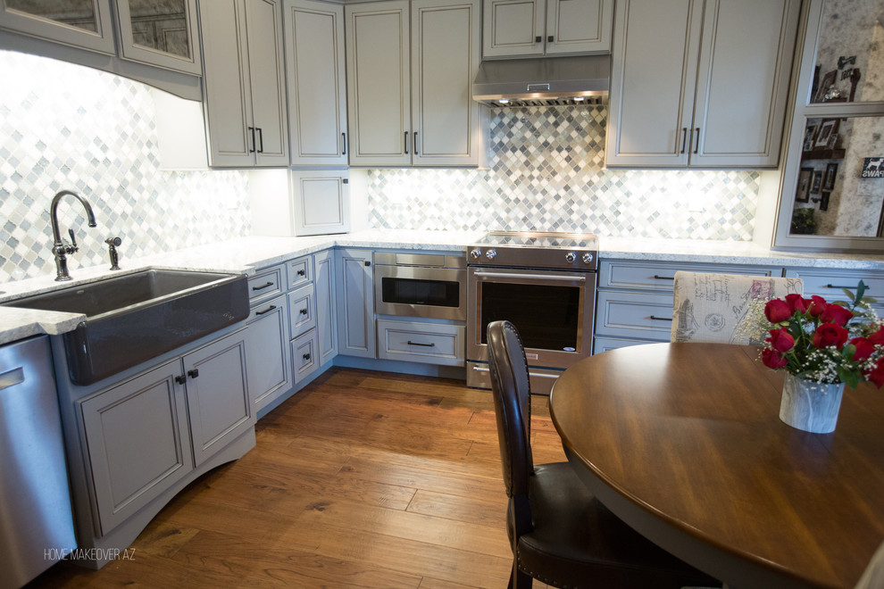 Example of a mid-sized classic l-shaped light wood floor and brown floor open concept kitchen design in Phoenix with a farmhouse sink, glass-front cabinets, gray cabinets, granite countertops, gray backsplash, limestone backsplash, stainless steel appliances and no island