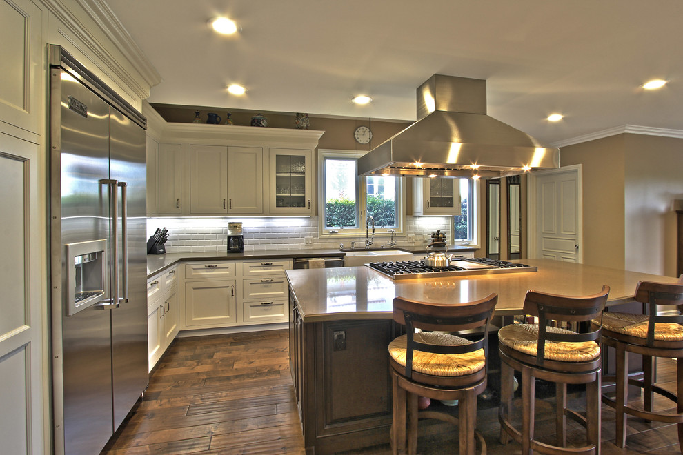 Example of a large classic l-shaped medium tone wood floor and beige floor eat-in kitchen design in Los Angeles with a farmhouse sink, shaker cabinets, white cabinets, quartz countertops, white backsplash, subway tile backsplash, stainless steel appliances, an island and brown countertops