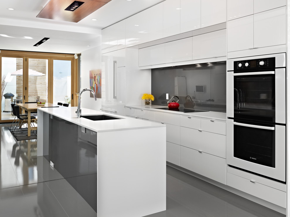 Idée de décoration pour une cuisine américaine parallèle minimaliste avec un évier encastré, un placard à porte plane, des portes de placard blanches, une crédence grise, un électroménager blanc, une crédence en feuille de verre et un sol gris.