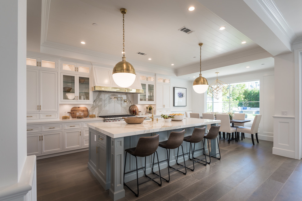 This is an example of a large traditional galley kitchen/diner in Los Angeles with a belfast sink, shaker cabinets, white splashback, stainless steel appliances, an island, brown floors, white cabinets and dark hardwood flooring.