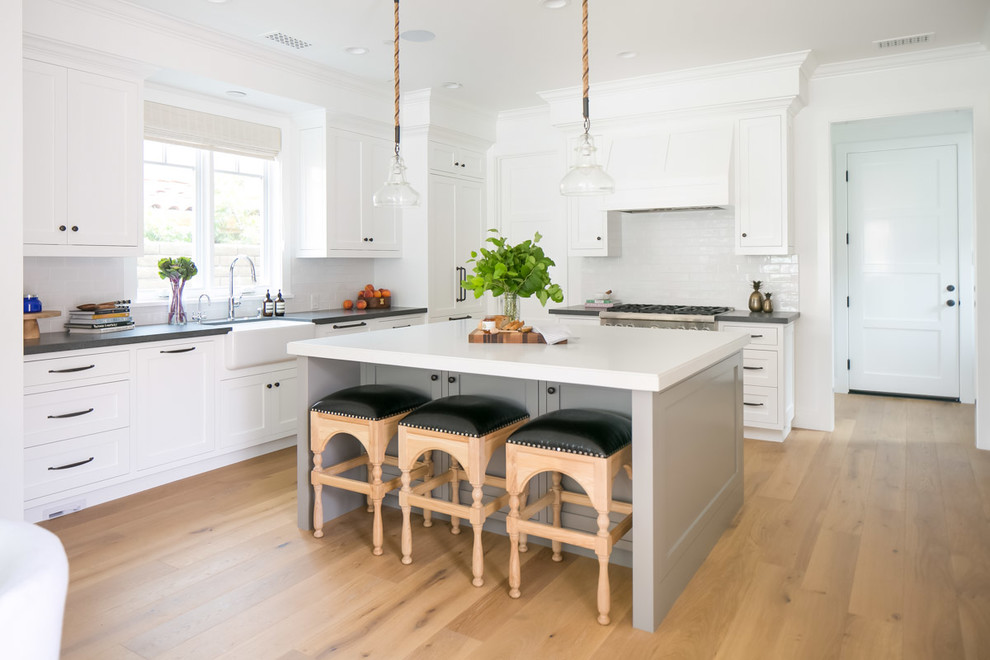 Photo of a medium sized coastal l-shaped kitchen/diner in Orange County with light hardwood flooring, a belfast sink, shaker cabinets, white cabinets, white splashback, stainless steel appliances, an island and beige floors.