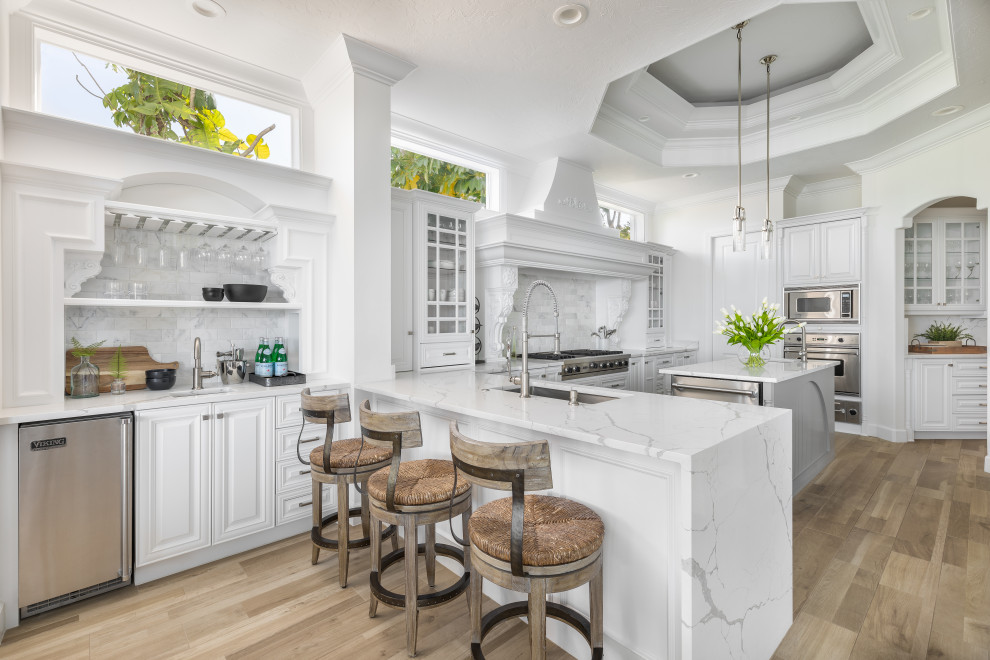 Tuscan u-shaped medium tone wood floor, brown floor and tray ceiling kitchen photo in Tampa with an undermount sink, raised-panel cabinets, white cabinets, white backsplash, stainless steel appliances, an island and white countertops