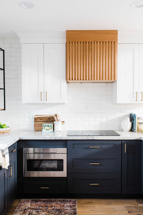 Premium RTA cabinets Canada: two-tone white-navy blue shaker cabinet, brass handles; slatted wood hood, white painted brick walls, quartz countertop and built-in microwave.