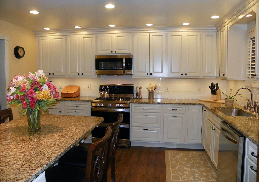Foto på ett avskilt, mellanstort vintage l-kök, med en undermonterad diskho, luckor med upphöjd panel, vita skåp, granitbänkskiva, vitt stänkskydd, stänkskydd i porslinskakel, rostfria vitvaror, mellanmörkt trägolv och en halv köksö