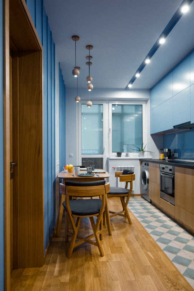 Inspiration pour une cuisine américaine design avec un placard à porte plane, des portes de placard bleues, une crédence bleue, une crédence en feuille de verre, un électroménager en acier inoxydable, aucun îlot, un sol multicolore et plan de travail noir.