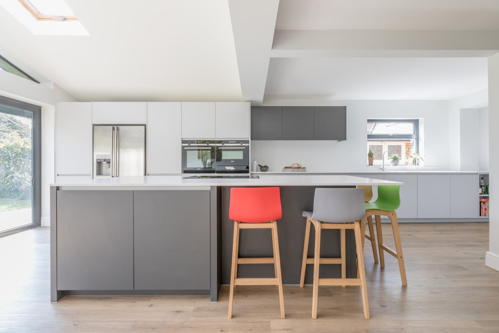 This is an example of a medium sized contemporary galley open plan kitchen in Sussex with an integrated sink, flat-panel cabinets, grey cabinets, composite countertops, stainless steel appliances, an island and white worktops.