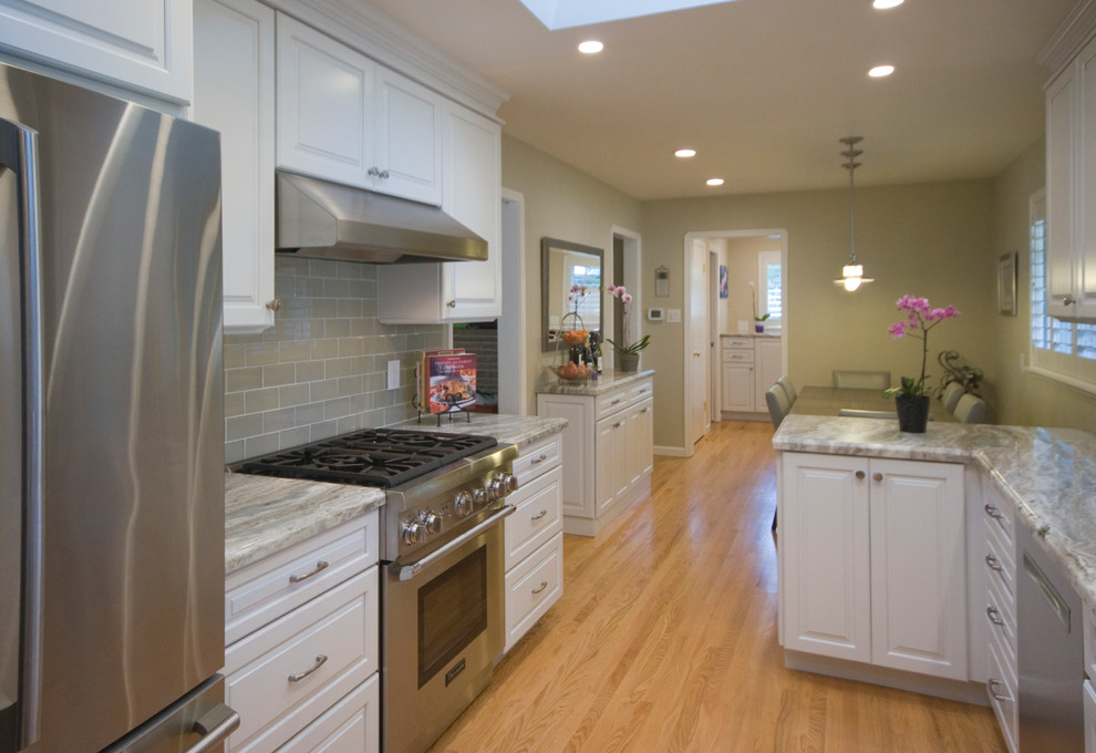 Enclosed kitchen - mid-sized traditional galley medium tone wood floor enclosed kitchen idea in San Francisco with an undermount sink, raised-panel cabinets, white cabinets, quartz countertops, gray backsplash, glass tile backsplash, stainless steel appliances and a peninsula