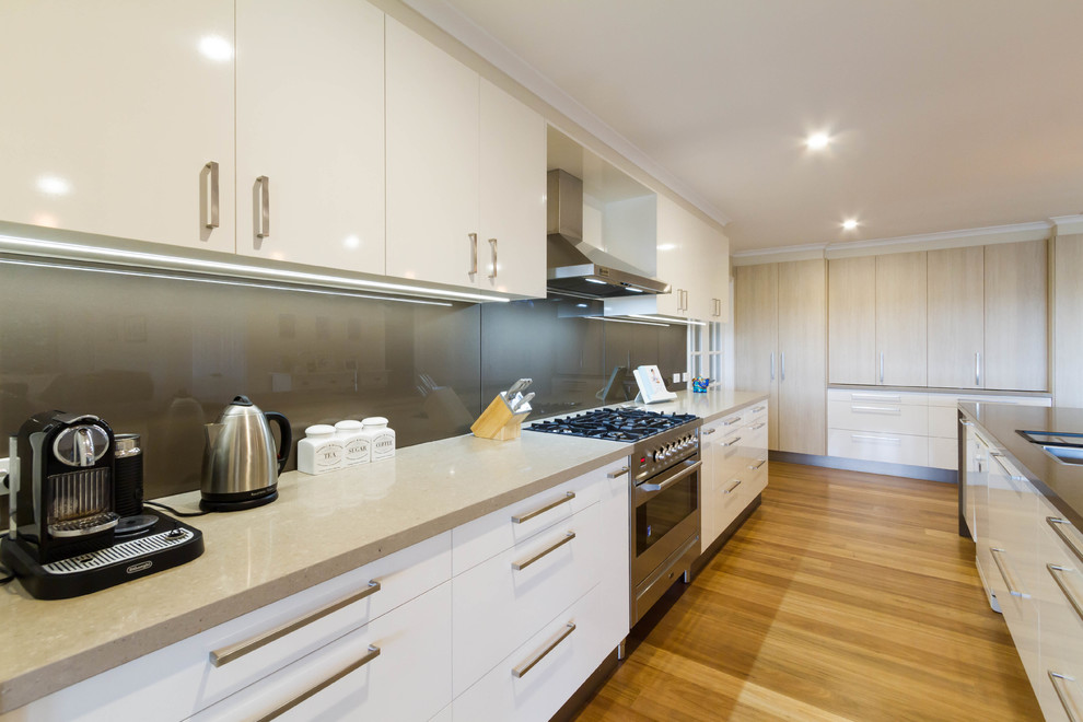 Photo of a large modern u-shaped kitchen pantry in Melbourne with a built-in sink, flat-panel cabinets, white cabinets, engineered stone countertops, grey splashback, glass sheet splashback, stainless steel appliances, medium hardwood flooring, an island and brown floors.