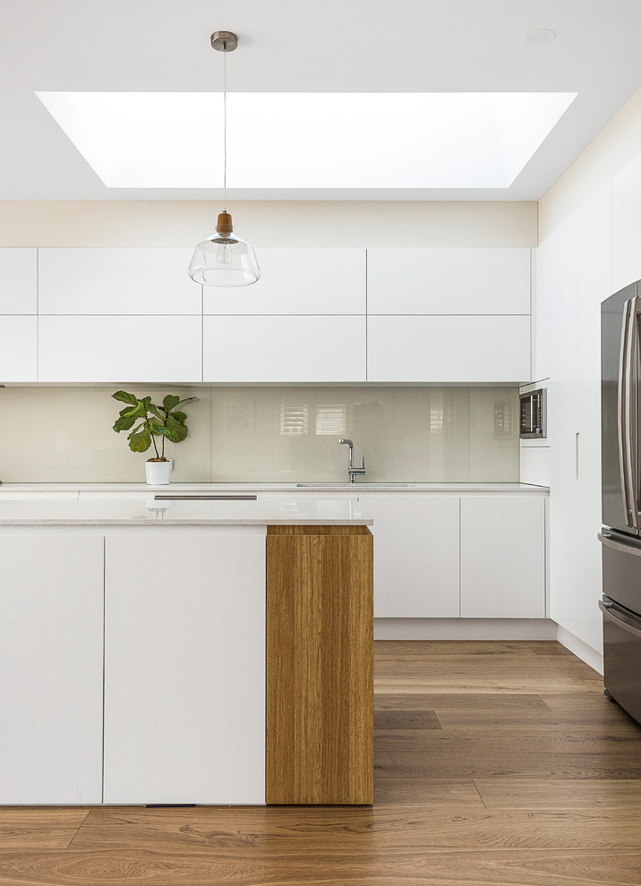 Minimalistisk inredning av ett stort vit vitt kök, med en undermonterad diskho, släta luckor, vita skåp, bänkskiva i kvarts, vitt stänkskydd, glaspanel som stänkskydd, rostfria vitvaror, mellanmörkt trägolv, en köksö och brunt golv