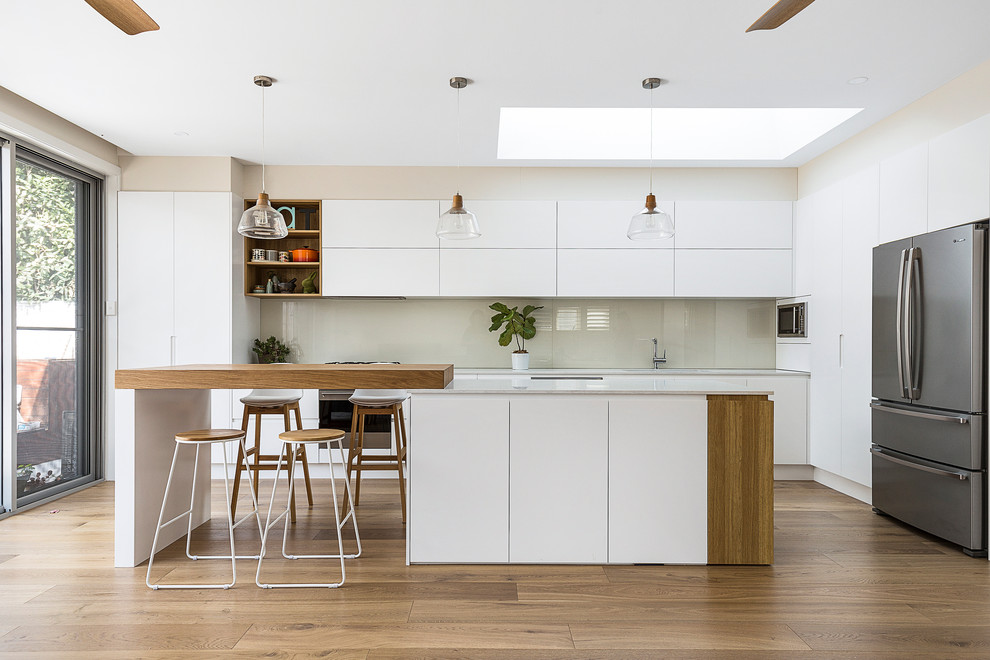 Foto på ett stort funkis vit kök, med en undermonterad diskho, släta luckor, vita skåp, glaspanel som stänkskydd, rostfria vitvaror, mellanmörkt trägolv, en köksö, bänkskiva i kvarts, vitt stänkskydd och brunt golv