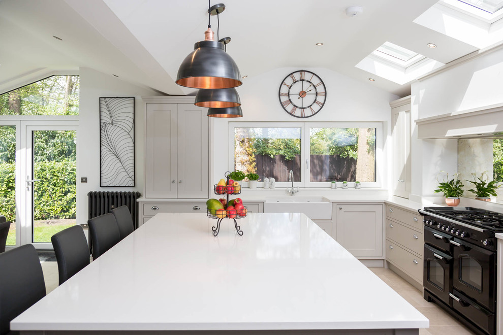 Traditional l-shaped kitchen/diner in Berkshire with a belfast sink, shaker cabinets, mirror splashback, black appliances, an island, beige floors and white worktops.