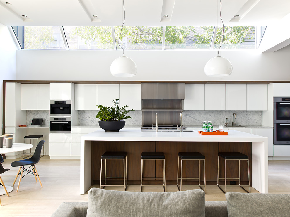 This is an example of a contemporary galley open plan kitchen in Chicago with a triple-bowl sink, flat-panel cabinets, white cabinets, grey splashback, stone slab splashback, stainless steel appliances, light hardwood flooring, an island, beige floors and grey worktops.