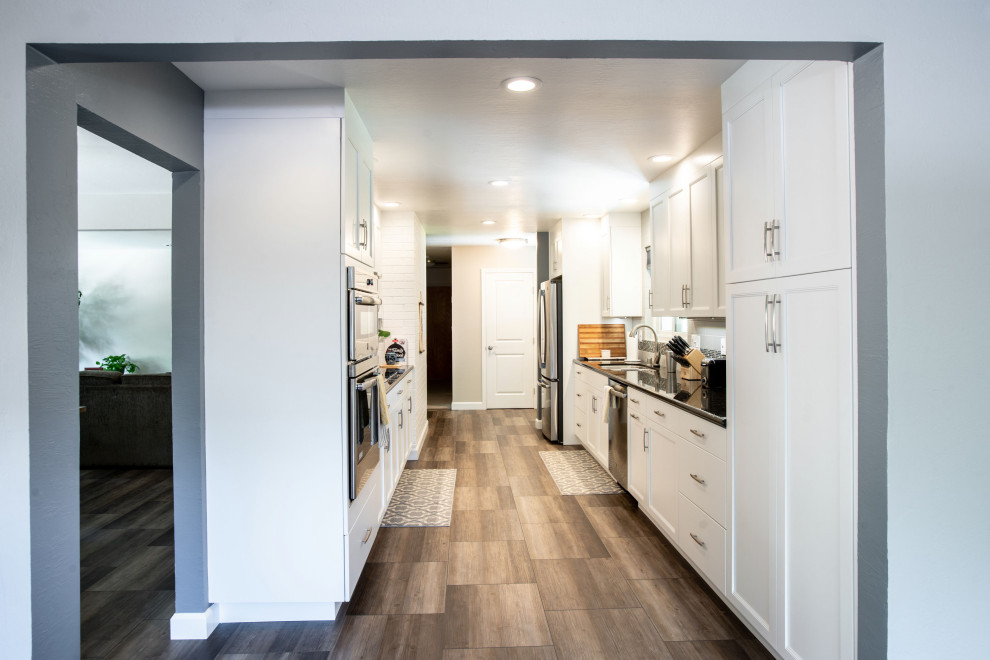 サンフランシスコにある小さなコンテンポラリースタイルのおしゃれなII型キッチン (ダブルシンク、落し込みパネル扉のキャビネット、白いキャビネット、グレーのキッチンパネル、サブウェイタイルのキッチンパネル、シルバーの調理設備、アイランドなし、茶色い床、黒いキッチンカウンター) の写真